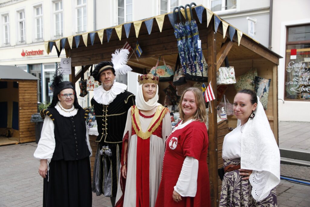 Fem glada personer framför en svensk monter på marknad iförda historiska kläder.